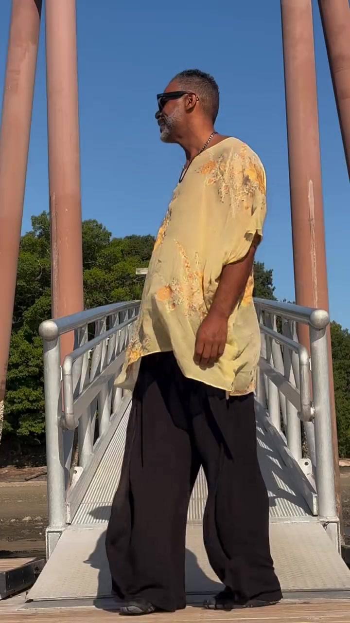 Kristopher in a handmade pale yellow top and wide dark trousers, standing on a wooden bridge.