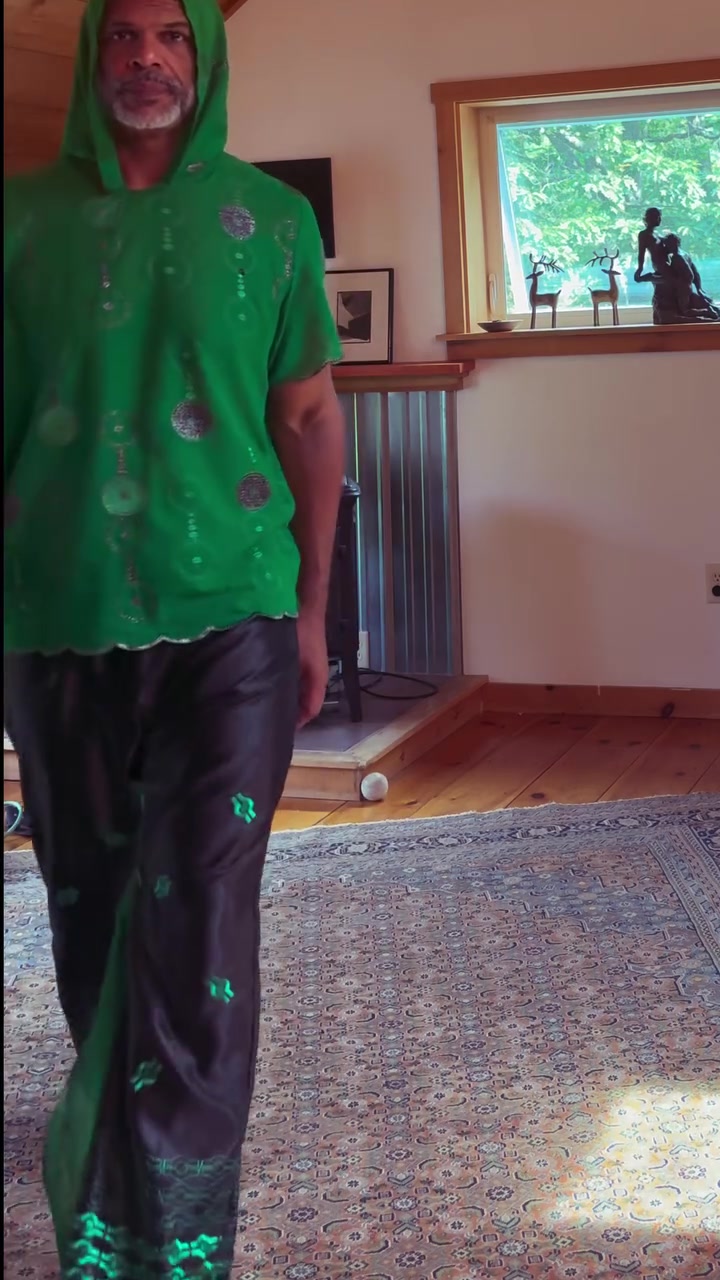 Kristopher wearing a handmade green hooded garment with patterned dark trousers.
